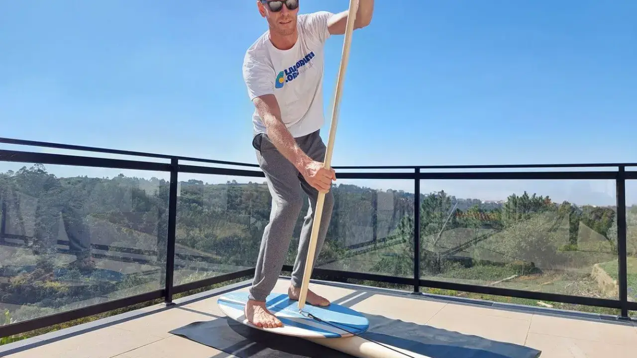 Mann übt Balance-Board-Training zuhause auf einer Terrasse. Er hält einen Paddelstock und balanciert auf einem Brett.