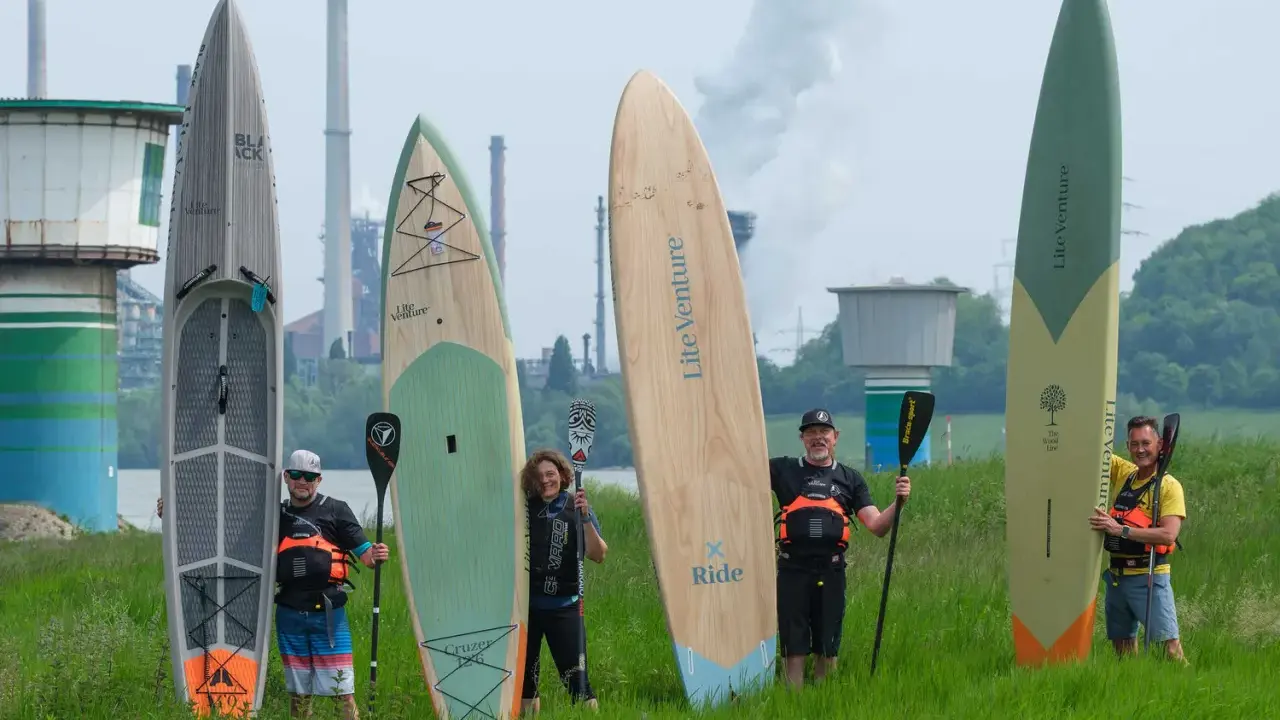 Fünf Personen mit SUP-Boards und Paddeln vor einer Industrieanlage. Die Boards zeigen fortgeschrittene SUP-Technik.