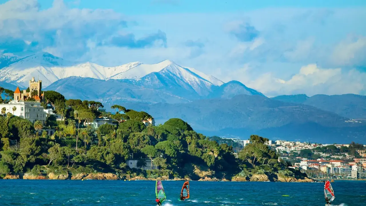 Windsurfen an der französischen Küste mit schneebedeckten Bergen im Hintergrund. Ein Schloss thront auf einem Hügel.