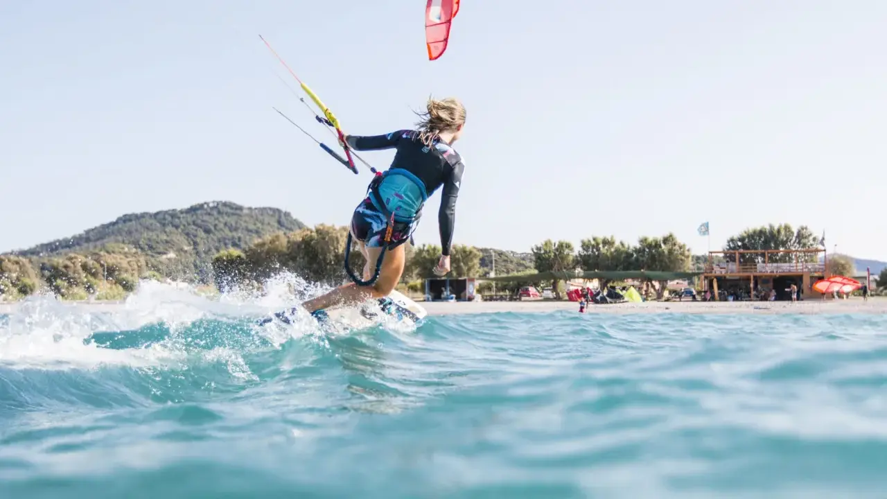 Eine Frau surft mit einem Kite auf Kos. Sie fährt auf einer Welle, Gischt spritzt. Im Hintergrund ist der Strand mit Bäumen und einem Gebäude zu sehen.