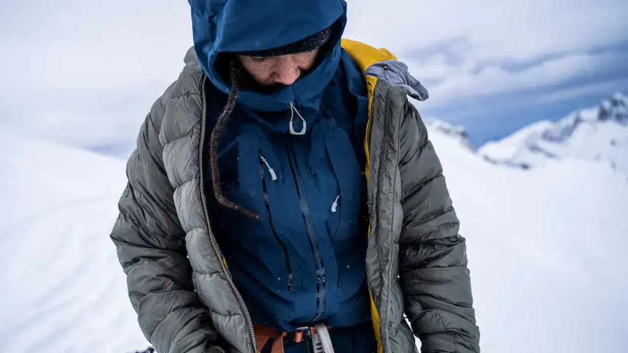 Person in Daunenjacke im Schnee. Die Jacke ist grau und blau, mit gelben Akzenten. Perfekt für das Daunenjacke waschen nach einer Bergtour.