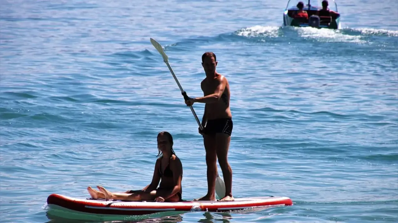 Ein Mann steht auf einem Stand Up Paddle Board auf dem Meer, während eine Frau sitzt. Im Hintergrund fährt ein Boot.