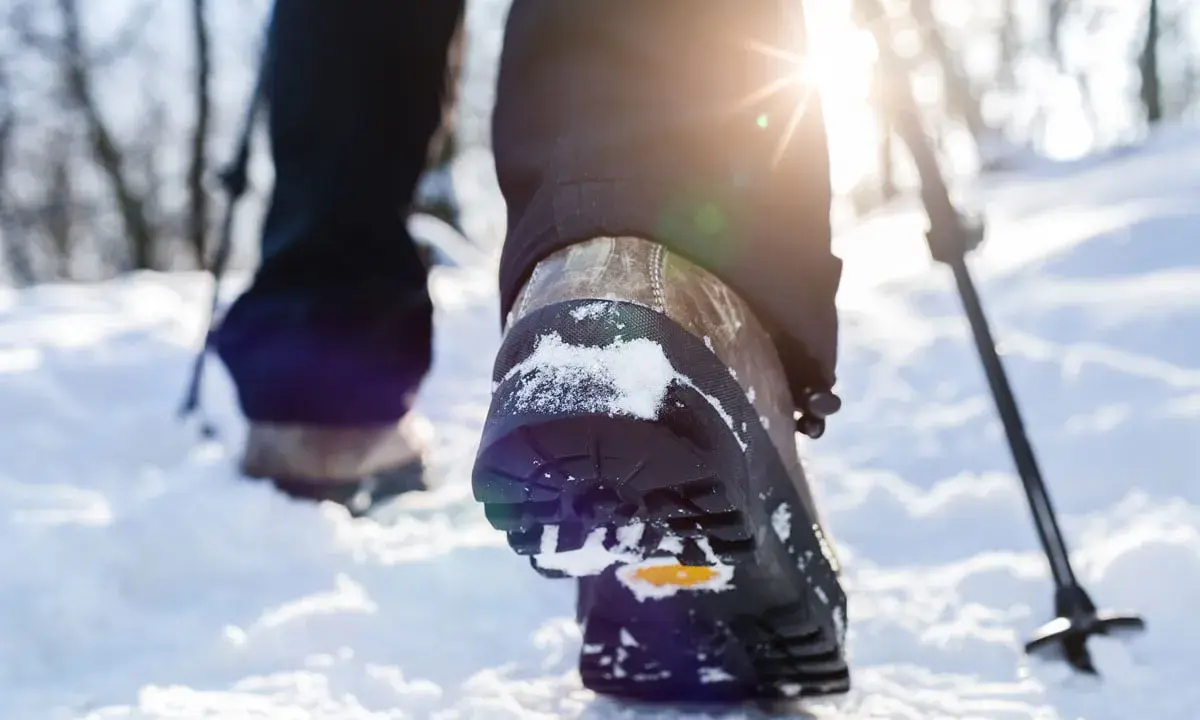 Schneeschuhwandern Schuhe mit Schnee bedeckt, während die Sonne durch die Bäume scheint. Ein Wanderstock ist im Hintergrund zu sehen.