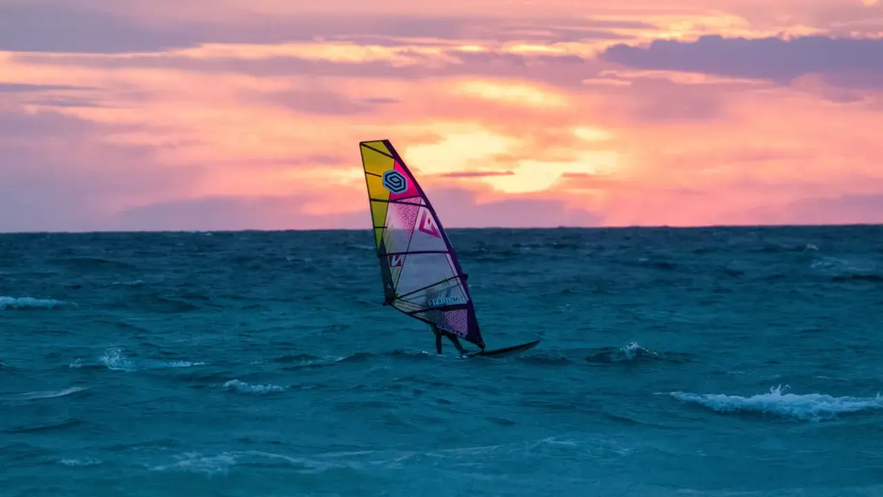 Ein Windsurfer gleitet bei Sonnenuntergang über das Meer. Die bunten Wolken spiegeln sich im Wasser. Ein perfekter Tag für Windsurfen Mallorca.