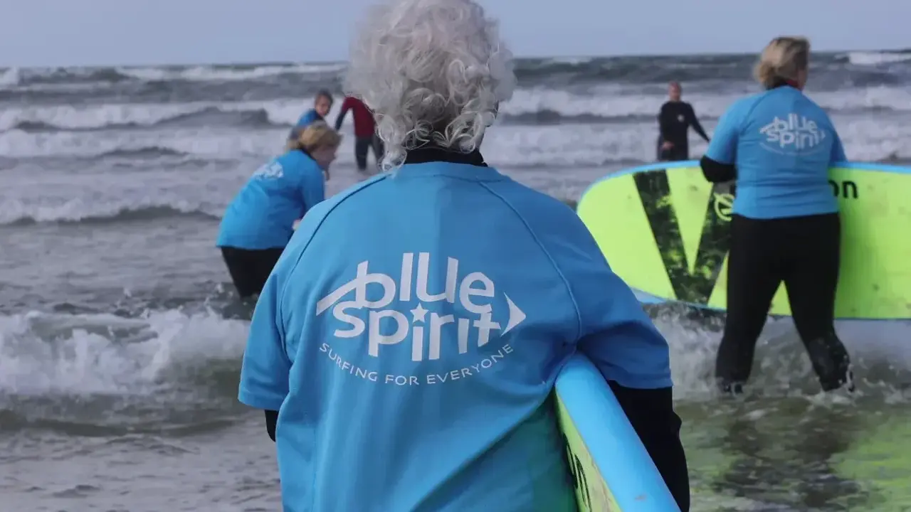Ältere Dame mit grauem Haar und Surfbrett, bereit für Hvide Sande Surfen. Mehrere Personen in blauen Shirts mit "Blue Spirit" Aufdruck im Wasser.