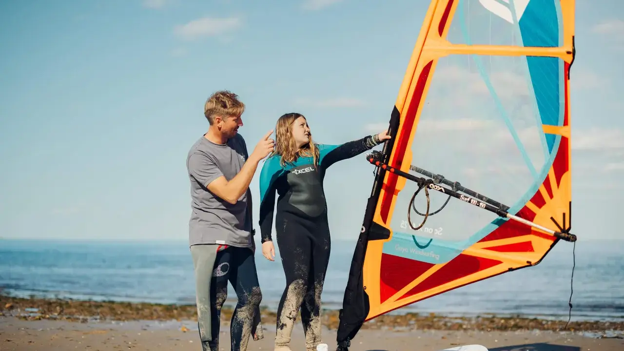 Vater erklärt Tochter das Windsurfen NRW. Sie stehen am Strand mit einem Segel und Board.