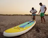 Zwei Männer bereiten ein SUP-Board am Strand vor. Einer pumpt es auf, fragt sich: "Sup, wieviel Bar?