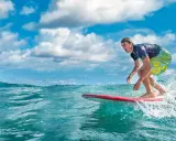 Ein Surfer reitet eine Welle vor der Küste der Dominikanischen Republik. Der Mann trägt ein blaues Shirt und bunte Badeshorts.