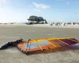 Windsurfen St. Peter-Ording: Segel und Brett liegen am Strand, bereit für den nächsten Ritt auf den Wellen. Strandkörbe und das markante Stelzenhaus im Hintergrund.