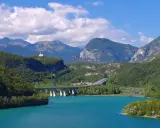 Türkises Wasser des Lago di Cavazzo umgeben von grünen Bergen. Eine Brücke überspannt den See, im Hintergrund eine kleine Kirche auf einem Hügel.