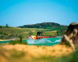Windsurfer springt über das Wasser am Lac de Ganguise. Grüne Hügel und blauer Himmel im Hintergrund.