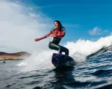 Frau surft auf einer Welle vor der Küste Lanzarotes, die Sonne scheint, blauer Himmel mit Wolken.