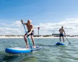 Zwei Männer genießen SUP auf der Nordsee. Sonnenschein, Strand und ein Leuchtturm im Hintergrund.
