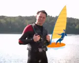 Mann in Neoprenanzug und Schwimmweste erklärt, wie funktioniert ein Neoprenanzug. Im Hintergrund eine stilisierte Windsurferin.