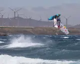 Windsurfer Britt Dunkerbeck springt hoch über den Wellen, im Hintergrund Windräder.