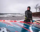 Mann in Neoprenanzug mit Windsurfbrett am Strand. Kaltes Wetter für Windsurfen in Deutschland.