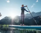 Eine Frau steht auf einem SUP-Board in Bayern, umgeben von Bergen und klarem Wasser.
