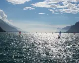 Windsurfer tummeln sich auf dem glitzernden Gardasee vor einer Bergkulisse.
