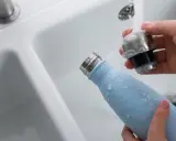 Hände reinigen eine hellblaue Trinkflasche unter fließendem Wasser im Waschbecken.