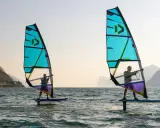 Zwei Personen beim Windsurfen in Italien. Die türkisfarbenen Segel sind im Gegenlicht zu sehen, im Hintergrund Berge.