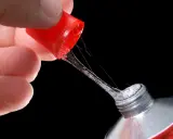 Ein Finger hält die rote Kappe eines Tubenklebers, aus dem zäher, glitzernder Kleber in Fäden heraustropft.