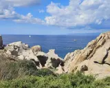 Felsformationen am Capo Testa mit Blick auf das blaue Meer und einen einsamen Segler unter bewölktem Himmel.