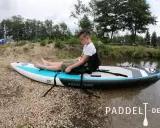 Junge sitzt auf SUP-Board, um den SUP Sitz zu befestigen. Das Board liegt am Ufer.