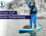 Person im blauen Neoprenanzug bereitet sich auf eine Winter SUP Tour vor. Die Landschaft ist mit Schnee bedeckt.