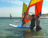 Zwei junge Männer bereiten sich auf das Windsurfen an der Ostsee vor. Ein weiterer Surfer ist in der Ferne zu sehen.
