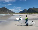 Zwei Surferinnen mit Neoprenanzügen und Surfbrettern am Strand in Norwegen. Die Berge im Hintergrund sind grün und die Wellen sind sanft.