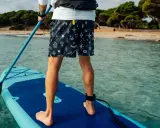 Person übt SUP Training auf einem Board im türkisfarbenen Wasser. Im Hintergrund eine sandige Bucht mit Bäumen.