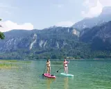 Zwei Frauen genießen Stand-Up-Paddling auf einem ruhigen See vor einer Bergkulisse. Perfekte sup spots für einen entspannten Tag.