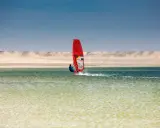Windsurfer mit rotem Segel gleitet über das Wasser bei Dakhla, Marokko, vor einer Wüstenlandschaft.