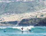 Surfer auf La Gomera reiten eine Welle, während im Hintergrund terrassierte Hänge und Häuser zu sehen sind.