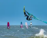 Windsurfer vollführt spektakulären Sprung über den Wellen. Mehrere andere Windsurfer sind auf dem Wasser zu sehen, was auf beliebte Windsurfing Spots hindeutet.