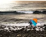 Ein Wingfoiler steht am steinigen Strand, bereit für den **shore break**. Die Sonne wirft ein warmes Licht auf die Wellen.