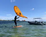 Person auf einem Board mit einem orangen Flügel, der über dem Wasser schwebt. Ein Schlauchboot liegt daneben. Foil surfen ohne Segel.