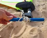 Hand hält blauen Griff, um das Segel zu spannen. Windsurf-Ausrüstung liegt im Sand.