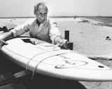 Jim Drake, ein Pionier des Surfens, lächelt stolz neben seinem Surfbrett am Strand.