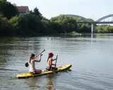 Zwei Mädchen genießen das Stand Up Paddling in Heidelberg. Im Hintergrund eine Brücke und Bäume.
