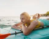 Junge Frau entspannt auf SUP-Board im Wasser. Ob Stand up paddling schwer ist, fragt sie sich lächelnd, während Freunde im Hintergrund planschen.