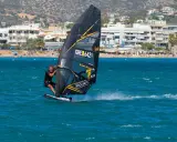 Ein Mann beim Windsurfen in Griechenland, mit Blick auf eine belebte Küste und Berge im Hintergrund.
