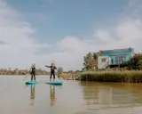 Zwei Frauen auf SUP-Boards am Makkum Strand, mit einem modernen Haus im Hintergrund.