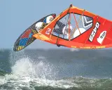 Lina Erpenstein fliegt mit ihrem Windsurf-Equipment über die Wellen. Das orangefarbene Segel und das bunte Board sind in der Luft.