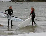 Zwei Mädchen bereiten sich auf das Windsurfen am IJsselmeer vor, tragen Neoprenanzüge und halten ihre Ausrüstung.