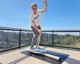 Mann übt Balance-Board-Training zuhause auf einer Terrasse. Er hält einen Paddelstock und balanciert auf einem Brett.