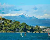 Windsurfen an der französischen Küste mit schneebedeckten Bergen im Hintergrund. Ein Schloss thront auf einem Hügel.