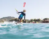 Eine Frau surft mit einem Kite auf Kos. Sie fährt auf einer Welle, Gischt spritzt. Im Hintergrund ist der Strand mit Bäumen und einem Gebäude zu sehen.