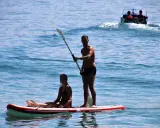 Ein Mann steht auf einem Stand Up Paddle Board auf dem Meer, während eine Frau sitzt. Im Hintergrund fährt ein Boot.