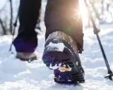 Schneeschuhwandern Schuhe mit Schnee bedeckt, während die Sonne durch die Bäume scheint. Ein Wanderstock ist im Hintergrund zu sehen.